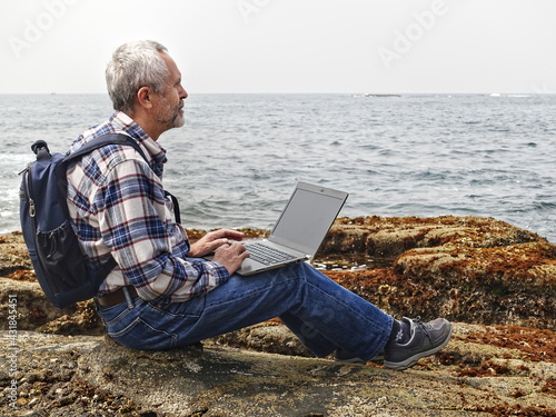 gray-haired man in jeans and a plaid shirt works with a laptop on the ocean shore