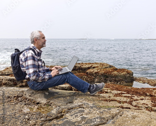 gray-haired man in jeans and a plaid shirt works with a laptop on the ocean shore