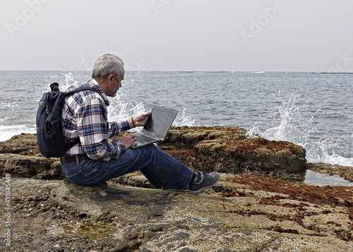 gray-haired man in jeans and a plaid shirt works with a laptop on the ocean shore