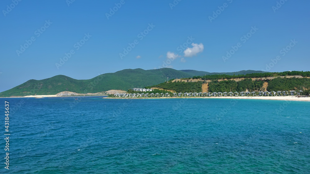 Summer seascape. There are ripples in the turquoise water. A row of villas can be seen on the sandy beach. Green mountains against the blue sky. Vietnam.