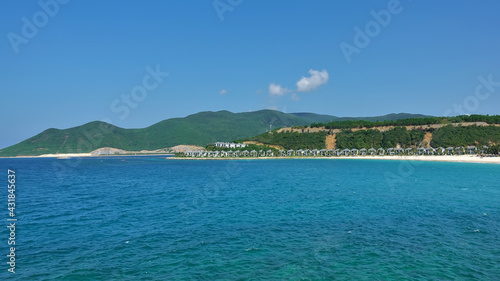 Summer seascape. There are ripples in the turquoise water. A row of villas can be seen on the sandy beach. Green mountains against the blue sky. Vietnam.