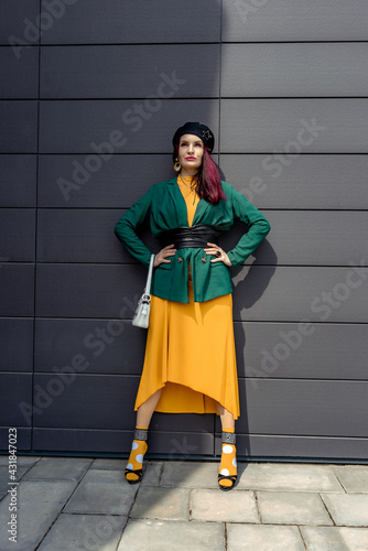Young woman in bright clothes, yellow skirt and green jacket. Yellow socks in sandals, beret on the head, hair with the color of magenta. Caucasian female fashion model standing against gray wall
