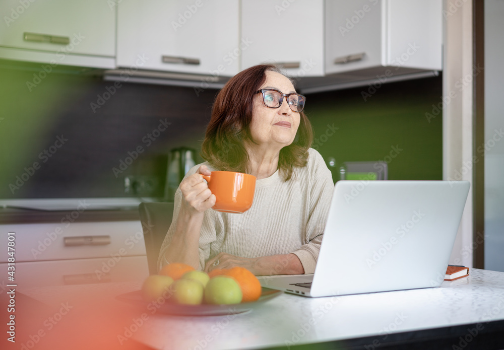 A senior woman in glasses working at home with a laptop and drinking coffee from a big mug sitting in the kitchen. A senior woman in glasses working at home with a laptop and drinking coffee from a big mug sitting in the kitchen.