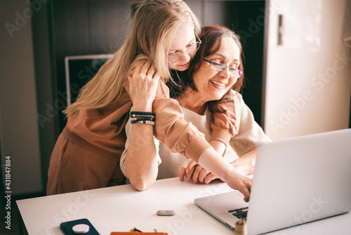 Beautiful happy grandmother with granddaughter in glasses spend time together at home looking at laptop photo