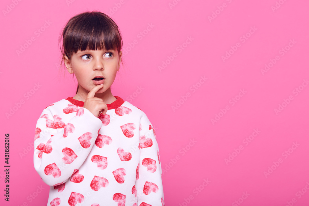 Surprised little girl looking to the side with her mouth wide open and her finger near mouth, copy space for advertising, wearing fashionable sweater, isolated over pink background. Surprised little girl looking to the side with her mouth wide open and her finger near mouth, copy space for advertising, wearing fashionable sweater, isolated over pink background.