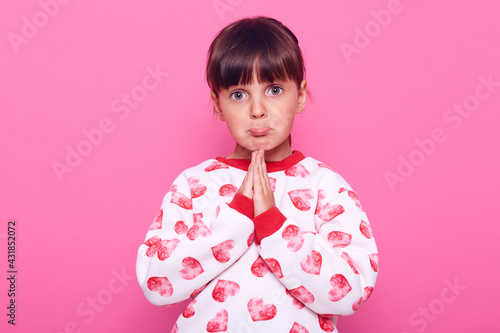Принти на полотні Sweet little girl with dark hair looks at camera with a pleading expression on her face, keeping palms together, praying, wearing a stylish sweater, isolated over pink background