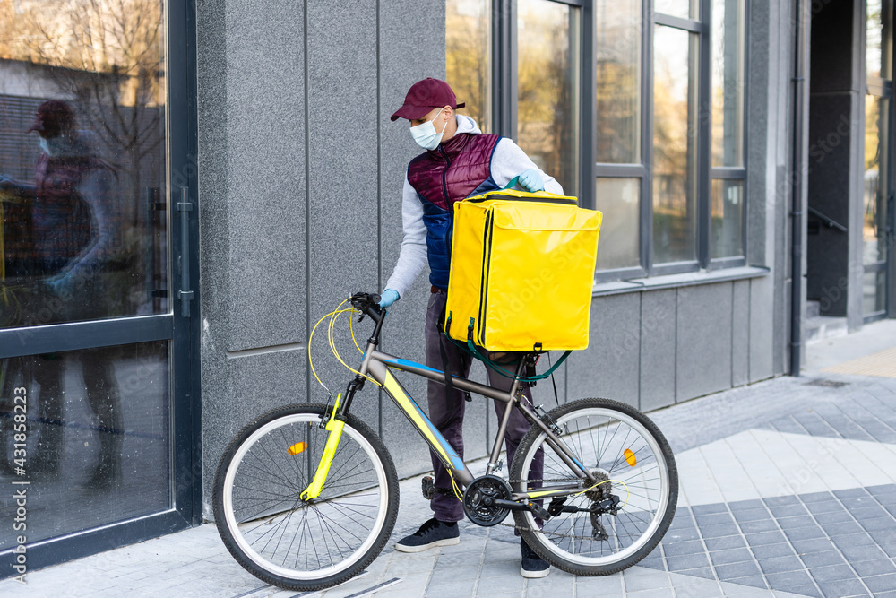 Young courier in medical mask delivering food with yellow thermal backpack, riding a bicycle in the city. Food delivery service concept