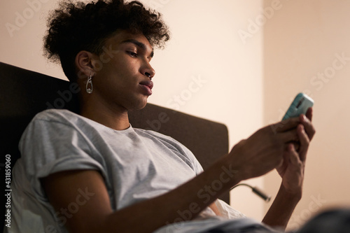 Social networks. Portrait of young guy with piercing using smartphone in his bed at night before going to sleep