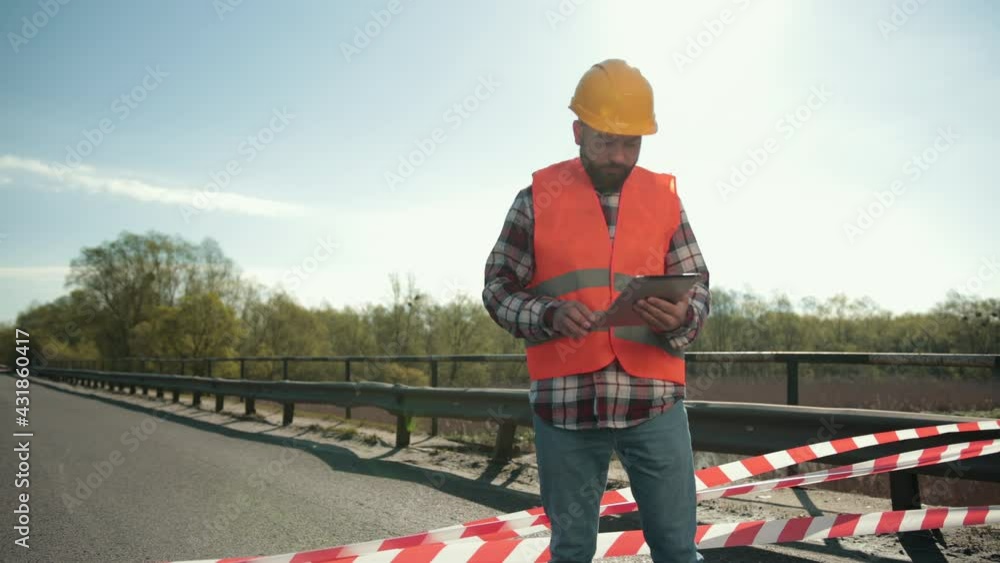 A young bearded man inspector in an orange vest and protective helmet with a tablet in his hand inspects a road accident, writes down a report A young bearded man inspector in an orange vest and protective helmet with a tablet in his hand inspects a road accident, writes down a report
