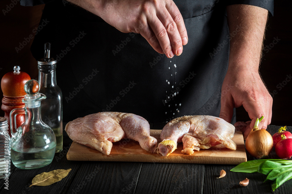 Сook or chef adds salt to the raw chicken legs. Preparation for grilling with chicken leg in the kitchen of a restaurant or cafe on a table with vegetables and spices
