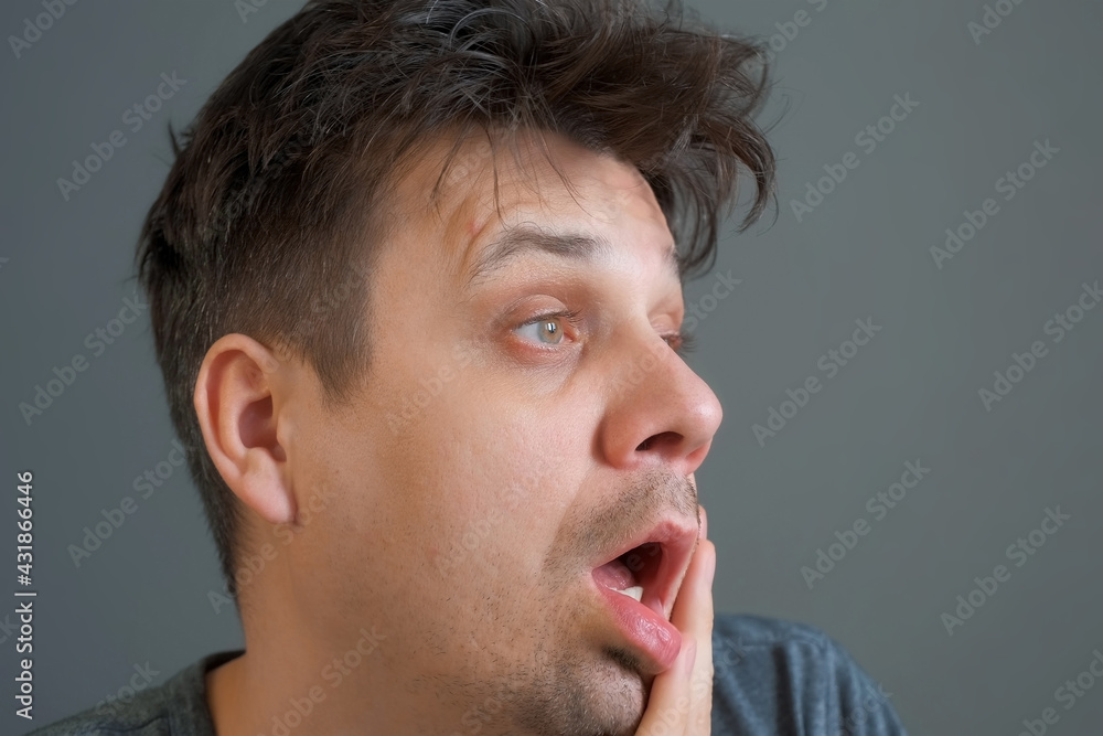 Obraz premium Tired boring shaggy young man on grey background. He is keeping his head by hands. Depression, apathy, and mental illness in the modern world. Middle age crisis.