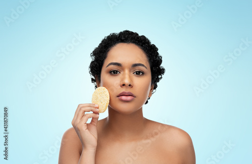 beauty, people and skincare concept - young african american woman with bare shoulders cleaning face with exfoliating sponge over blue background