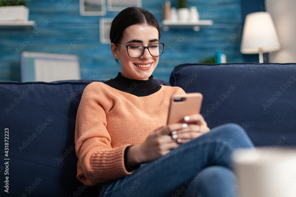 Relaxed student sitting on couch in living room while writing message holding modern smarphone in hands. Teenager woman chatting on social network looking at phone screen - obrazy, fototapety, plakaty 