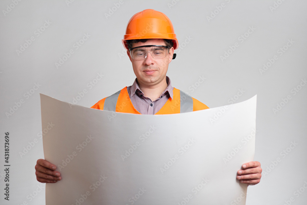 Man in builder's uniform looking in opened project, holding large sheet of paper on grey background. Construction worker in protective helmet on head and eyeglasses. Concept building industry