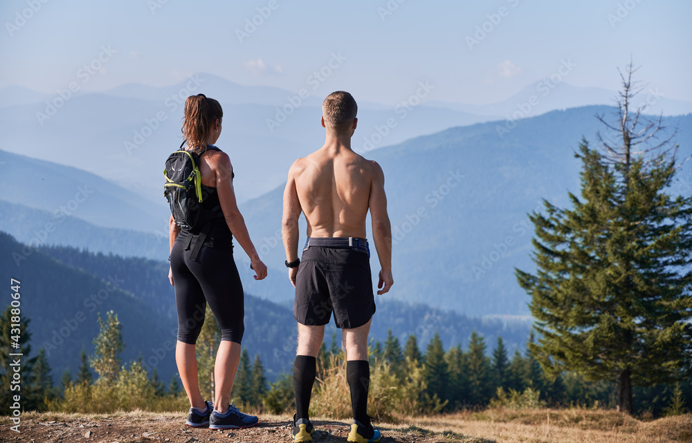 Obraz premium Back view of athletic young man and woman enjoying beautiful view of blue sky and mountains. Sporty couple standing on top of the hill and looking at majestic mountains. Concept of sport, travelling.