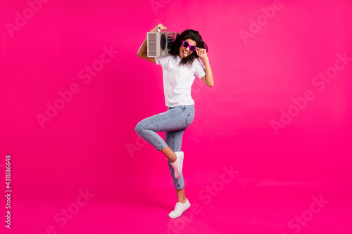 Full body profile photo of cool lady hold boom box tongue out wear spectacles t-shirt jeans sneakers isolated on pink background