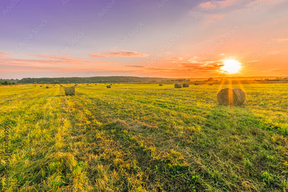 Obraz premium Scenic view at beautiful sunset in a green shiny field in village farm with hay stacks, cloudy sky, golden sun rays, anazing summer valley evening landscape