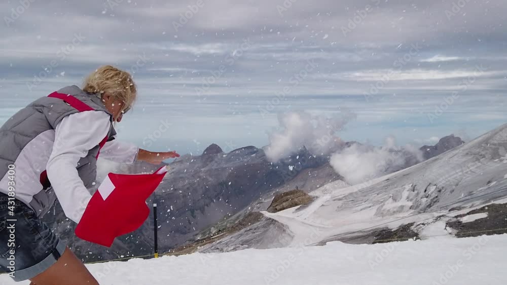 SLOW MOTION: Woman trekking in the snow storm with a Swiss flag. Top of Titlis glacier in the Uri Alps at 3028 meters. Cantons of Obwalden and Bern, Switzerland, Europe. Summer season. SLOW MOTION: Woman trekking in the snow storm with a Swiss flag. Top of Titlis glacier in the Uri Alps at 3028 meters. Cantons of Obwalden and Bern, Switzerland, Europe. Summer season.