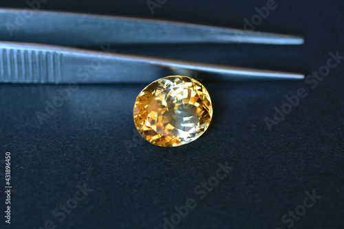Natural yellow uneven distributed color in crystal massif of oval faceted loose citrine gemstone setting for making jewelry. Dark background. Tweezers on the back. High gamma contrast. Gemology theme.