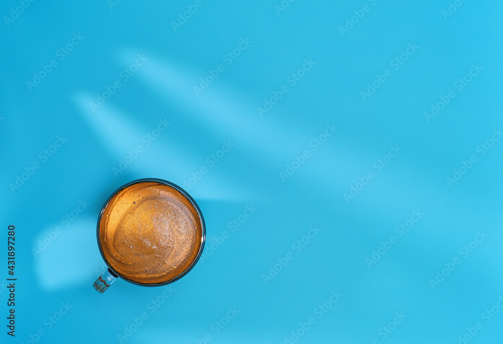 Coffee in glass cup is positioned against a bright blue background, shadows from leaves on the table. Strong espresso with aromatic crema, copy space layout. Banner with coffee for breakfast