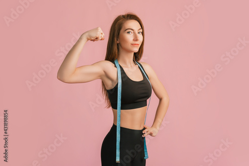 Fitness girl smiling and posing hold measure tape in black sportswear on a pink background. Slim woman with a beautiful athletic body and tanned skin © Daria Lukoiko