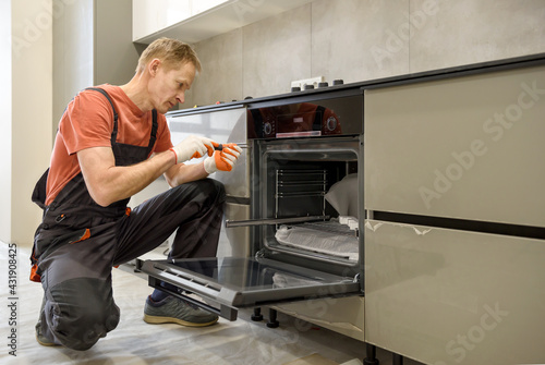 The worker is installing an electric oven in the kitchen furniture.