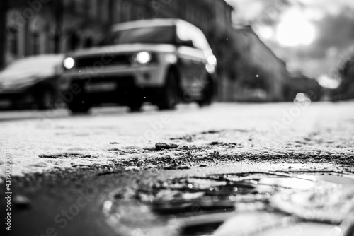 Bright winter sun in a big city, near a passing car in the snowfall. Close up view of a hatch at the level of the asphalt