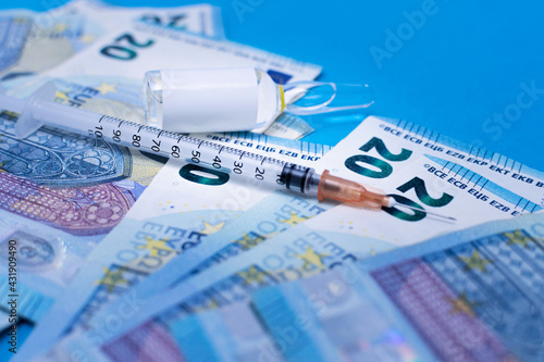 Syringe to take advantage of the coronavirus vaccine, the flu vaccine on money in 20 euro bills. Next to the vaccine vial part of the vial syringe and banknotes are out of focus on a blue surface.