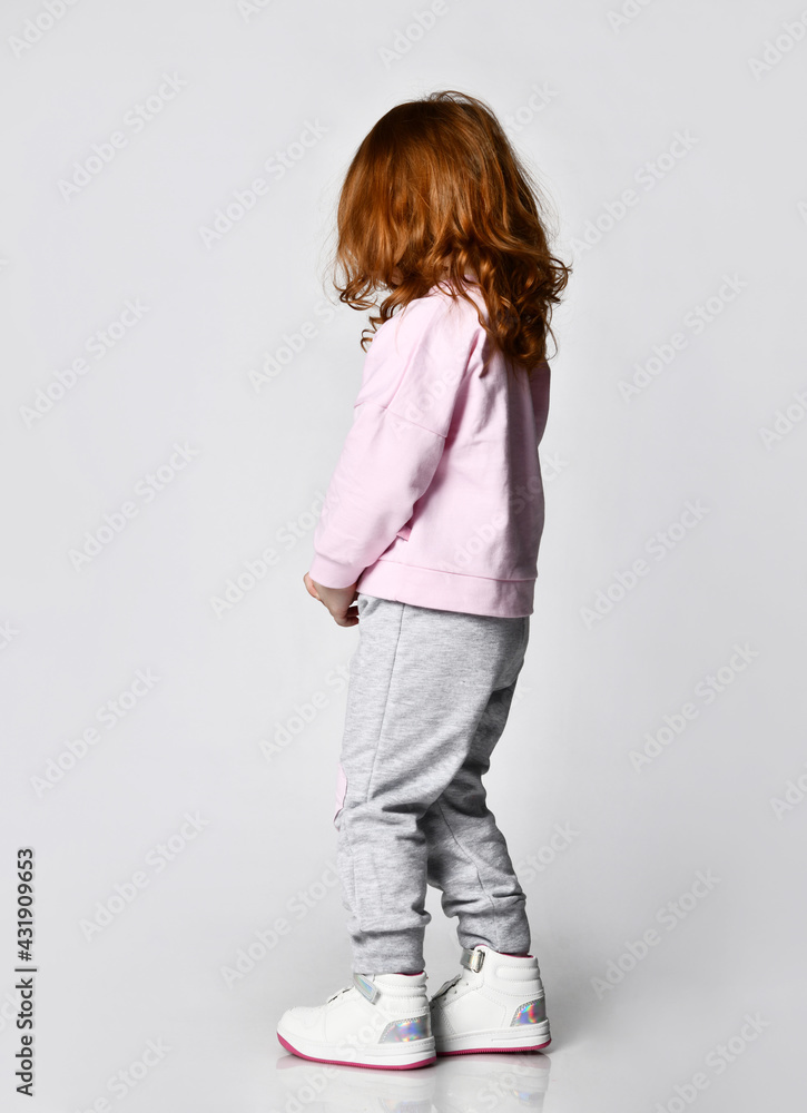 Rear view of a redhead girl in a pink sweatshirt walking towards the free copy space against a gray wall. Happy childhood and fashionable children's clothing