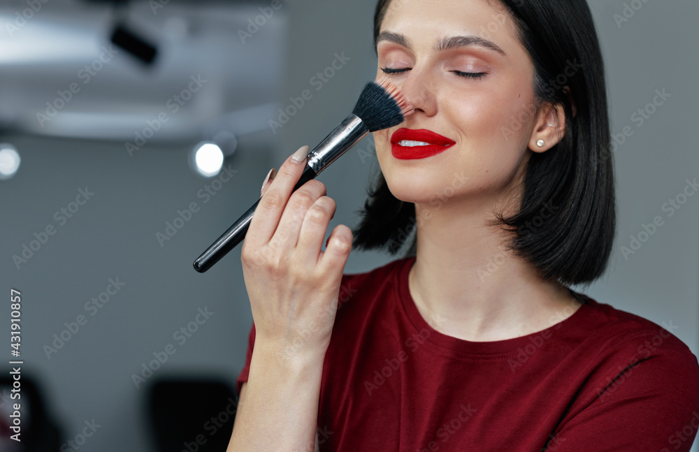 Positive young woman with red lips applying cosmetic powder with a brush on her nose, doing makeup herself. Positive young woman with red lips applying cosmetic powder with a brush on her nose, doing makeup herself.