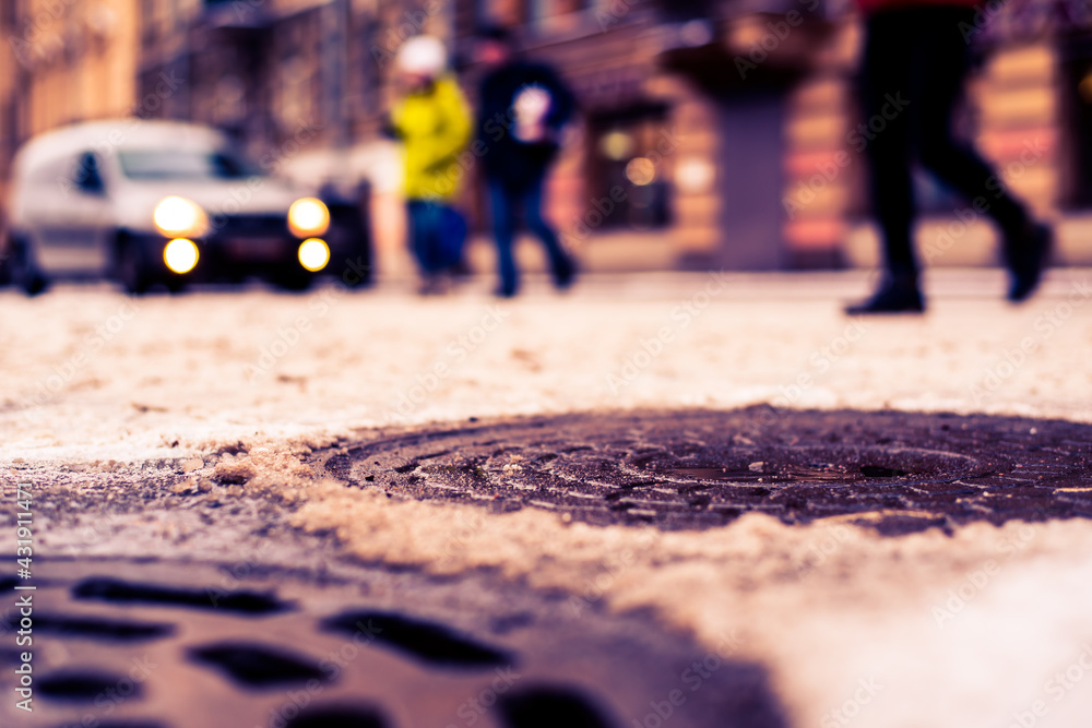 Obraz premium Snowy winter in the big city, pedestrians run across the road in front of riding car. Close up view of a hatches at the level of the asphalt
