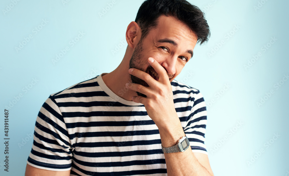 Happy young man laughing, covers his mouth with hand, looking to the camera, posing for advertisement, isolated on blue background. Handsome male has joyful expression, posing against white blue wall. Happy young man laughing, covers his mouth with hand, looking to the camera, posing for advertisement, isolated on blue background. Handsome male has joyful expression, posing against white blue wall.
