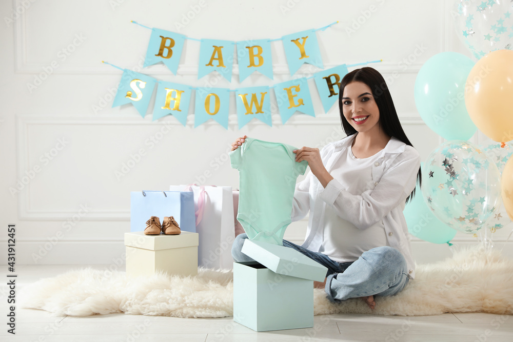Happy pregnant woman holding onesie in room decorated for baby shower party Happy pregnant woman holding onesie in room decorated for baby shower party