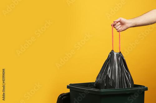Obraz na płótnie Man throwing garbage bag into bin on yellow background, closeup. Space for text