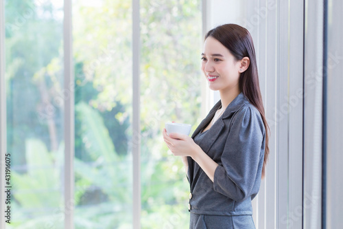 Asian working female wears grey dress stands and holds coffee cup in her hands in the morning at office room.