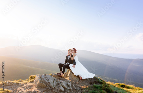 Front view of stylish guy and girl embracing sitting on a rock on a background of sky. Dating in the fresh air. Concept of outdoor love story