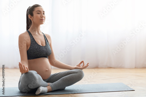 Beautiful pregnant woman sitting on yoga mat in living room, practicing meditation