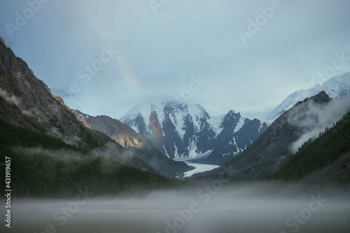 Atmospheric alpine landscape with mountain lake in fog and snowy mountains with rainbow in rainy weather. Gloomy scenery with mountain lake with rainy circles in fog on water and low clouds in valley.