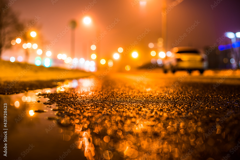 Foggy night in the big city, the street lighting lanterns and a parked car. Close up view from the level of the puddle on the pavement