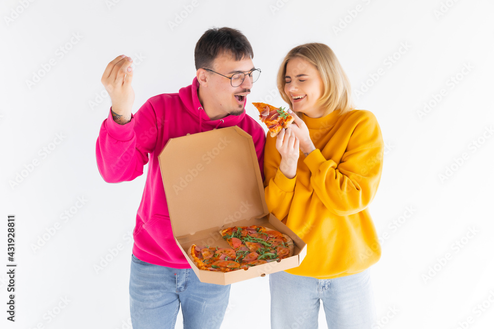 Portrait of cheery couple man and woman in colored sweaters smiling while eating pizza from box isolated over white background