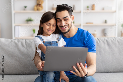 Smiling dad and daughter sitting on couch, using laptop