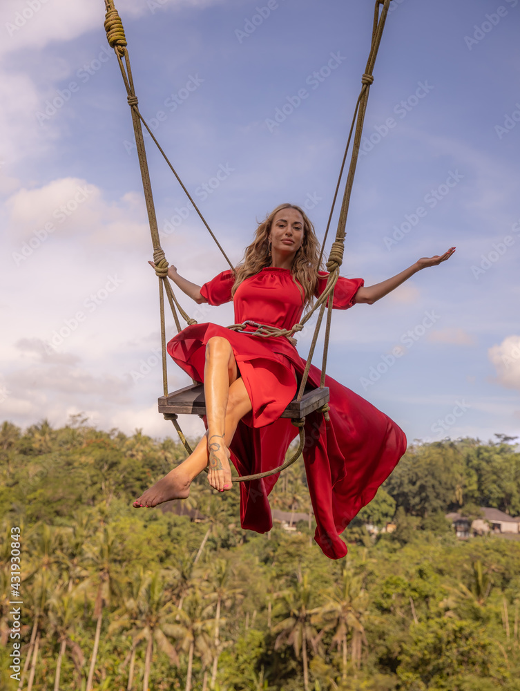 Bali swing trend. Caucasian woman in long red dress swinging in the jungle rainforest. Vacation in Asia. Travel lifestyle. Blue sky. Bongkasa, Bali, Indonesia