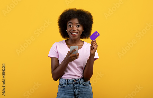 Easy money transfer. Positive black lady showing credit card and holding her mobile phone over yellow background