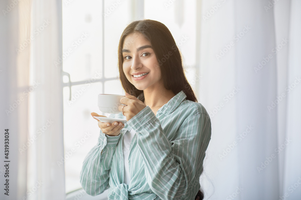 Lovely young Indian woman having coffee break in front of window at home Lovely young Indian woman having coffee break in front of window at home