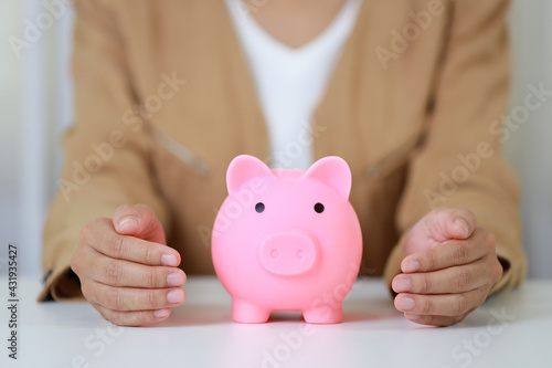 Young active business woman hands in casual dress sitting and protecting piggy bank on white table. Insurance and security concept.