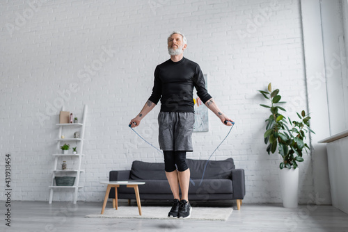 bearded man in sportswear exercising with jumping rope in modern living room.