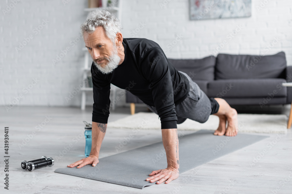 tattooed man with grey hair doing plank on fitness mat near dumbbells and sports bottle in living room.
