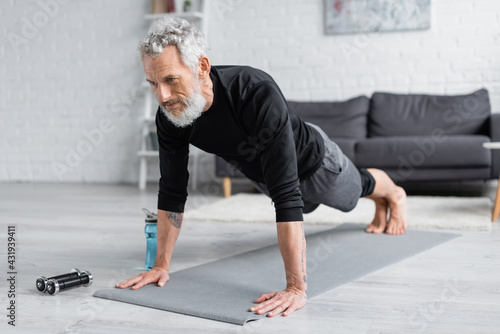 Принти на полотні tattooed man with grey hair doing plank on fitness mat near dumbbells and sports bottle in living room