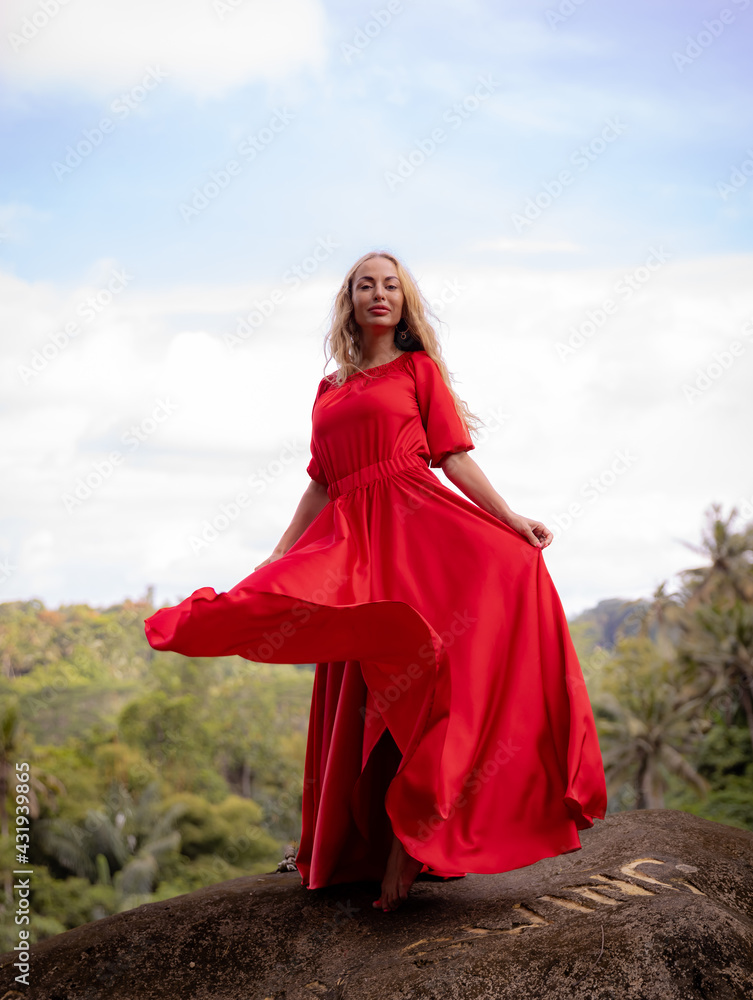 Naklejka premium Bali trend photo. Caucasian woman in long red dress standing on big stone in tropical rainforest. Vacation in Asia. Travel lifestyle. Breathtaking view. Bongkasa, Bali, Indonesia