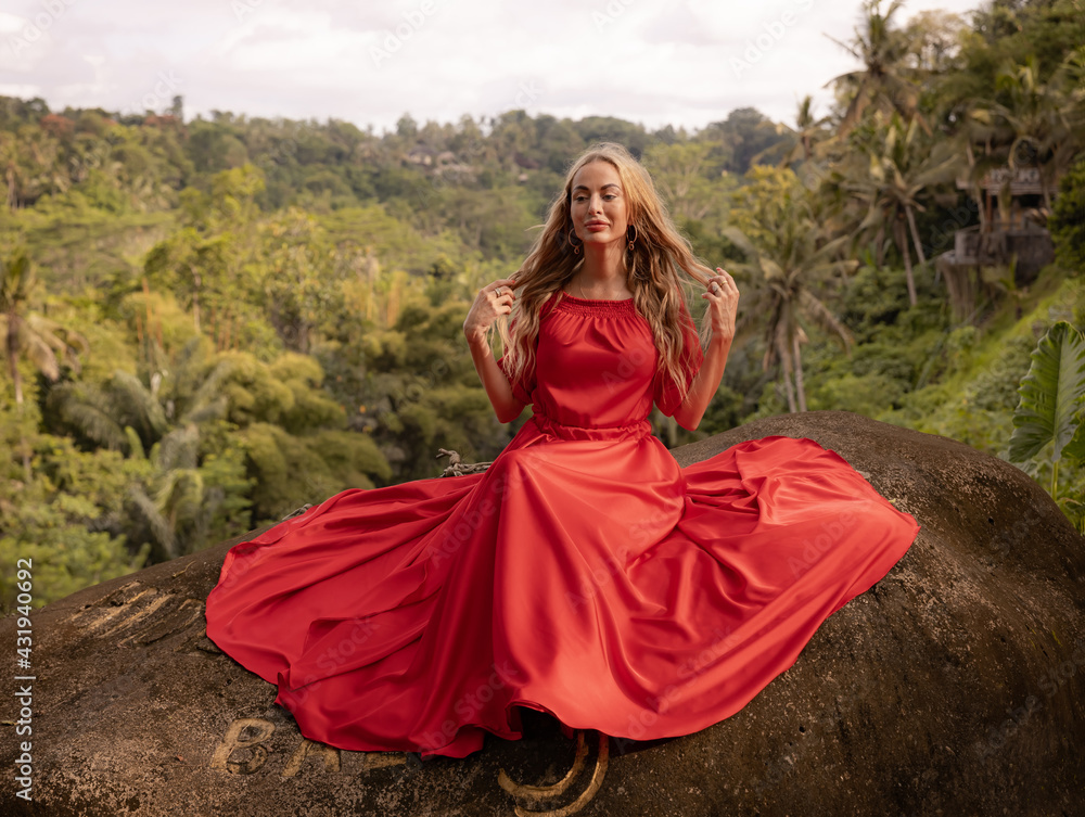 Bali trend photo. Caucasian woman in long red dress sitting on big stone in tropical rainforest. Vacation in Asia. Travel lifestyle. Breathtaking view. Bongkasa, Bali, Indonesia